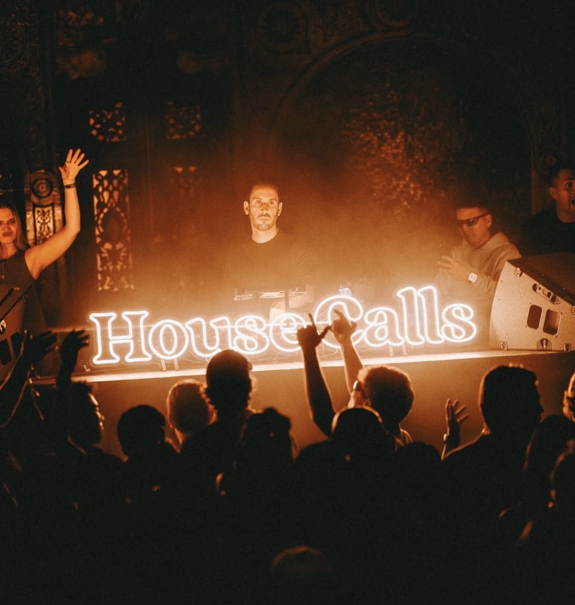 DJ performing behind the glowing House Calls sign with the audience in the foreground